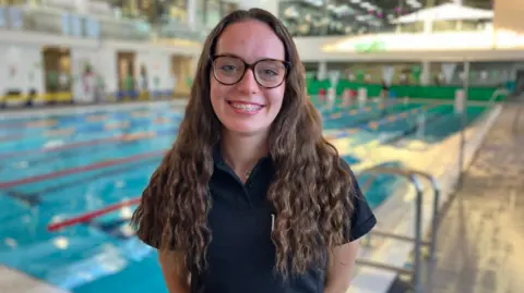BBC/Edward Sault Darcie Curtis with long wavy hair and glasses stands in front of an indoor swimming pool. She is wearing a dark collared shirt and is facing the camera. The background contains swimming lanes, pool railings, and a well-lit indoor aquatic facility with large windows and upper-level viewing areas, softly blurred to keep focus on Darcie.