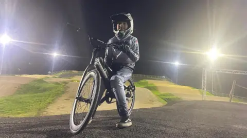 Ben Fisher Archie is posing at a track at night with floodlights on in the background. He is dressed in grey cycle wear with a grey helmet.