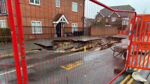 A sinkhole behind a red metal fence. There is a red brick house in the background, as well as some green trees. 