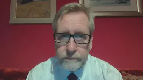 Donald Macaskill, a man with greying hair, a beard and glasses, sitting on a couch with red walls behind him. Paintings hang on the walls and he is wearing a white shirt with tie.