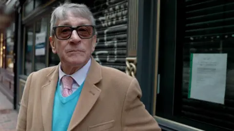 Ben Moore/BBC Man in glasses standing on a street looking into the camera. He is wearing a white shirt, pink tie, blue jumper and a beige coat. 