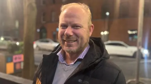 Nicky Crancher - a man with short blonde hair smiles at the camera. In the background are three cars waiting in traffic.