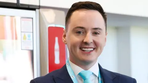 LDRS Darren Grimes is smiling and wearing a navy suit with a light blue tie and a Reform rosette in the same colour. His short brown hair is swept to the side. 