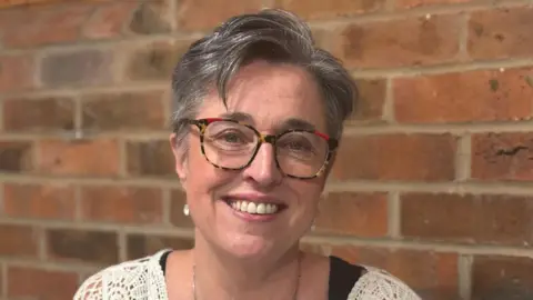 Gill Gray is smiling brightly at the camera. She has short grey hair and glasses.