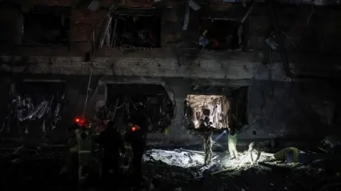 Reuters Emergency workers, dressed in yellow, work by torchlight as they inspect a crater from a missile attack at the base of a badly damaged building.