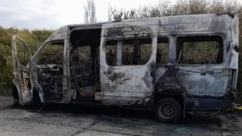 A burnt out minibus in front of a green hedge