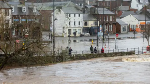 PA Media The Whitesands area of Dumfries is flooded by water from the River Nith moving up towards a row of businesses with a small number of people standing watching the scene