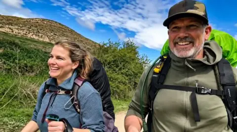 Supplied by Christie Mitchell An image showing Christie side by side with her partner Steve mid-hike. They are both smiling and wearing outdoorsy gear, including hiking backpacks. Behind them is a blue sky and grass-covered hills. 