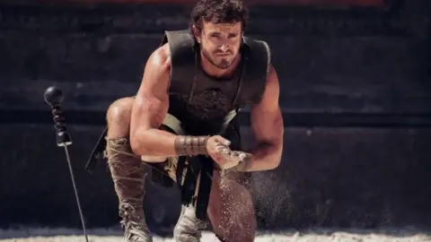 Paramount Paul Mescal plays Lucius, second Gladiator film. A man crouches, kneeling on the ground wearing a leather upper body sleeveless armour, calf length boots and rubs his hands together. Beside him, a sword embedded in the ground