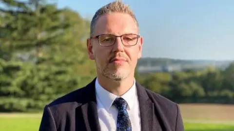 West Midlands Police and Crime Commissioner Police and Crime Commissioner John Campion looks straight ahead. He is wearing glasses a black jacket and white shirt 
