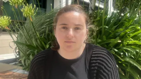 BBC Verity O'Connell is sat in Trenton Square with flowers behind her. She is wearing a black cardigan and a black top. She has brown hair and brown eyes.