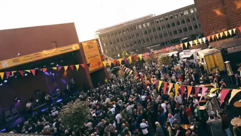 A large crowd is watching a band playing on a stage in front of the Fire Station venue in Sunderland. String of pink, yellow and red flags are hanging in the air.
