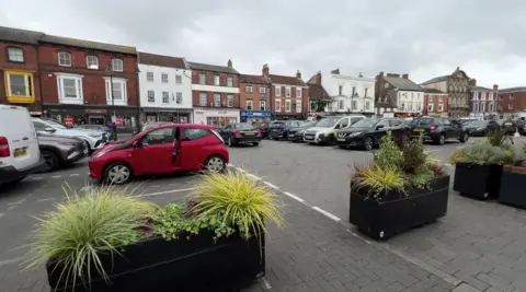 A view of a large market place, laid with grey block paving stones, with parking in the centre and black planters, topped with green shrubs, at the front of the photo. Cars are parked behind them. The square is surrounded by buildings, some Georgian-style, some more modern retail units.