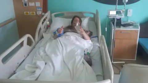 Supplied A woman prone in a hospital raises both thumbs. She is wearing an oxygen mask. 