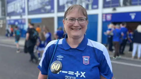 Aimee Dexter/BBC Lisa Fisher is standing in front of a football stadium. She has short light brown hair and is wearing a blue and white Ipswich Town football T-shirt. She has a badge pinned to one side of her T-shirt which says 60.