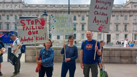 Action Against ASGSF Changes Three people all wearing blue hold placards above their heads. One reads 'stop failing our adopted kids' another reads 'betraying adopted children again'. In the background is a large stone building.