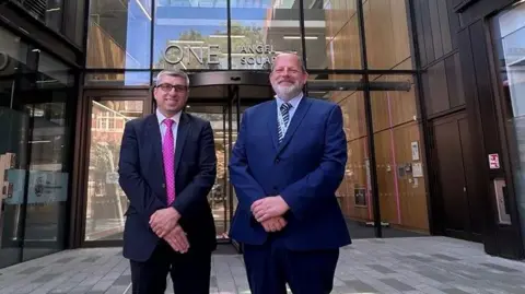 Reform UK Mark Arnull (left) stands in a dark suit and pink tie. James Petter stands beside him in a navy suit with a light blue shirt. Both are standing in front of One Angel Square, the glass-fronted headquarters of West Northamptonshire Council.
