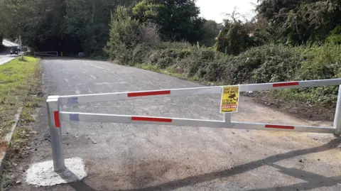 North Northamptonshire Council A lay-by with a red and white gate across it at either end. On the left is a verge and beyond it a road. On the right is a verge, hedge and trees. 