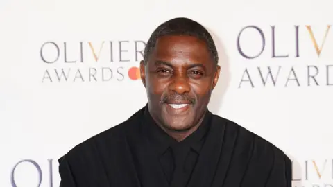 PA Media Idris Elba wearing a black suit with a black shirt and tie. He has very short dark hair and a stubbly black beard. He is stood in front of a white background with the graphics that read Olivier Awards.