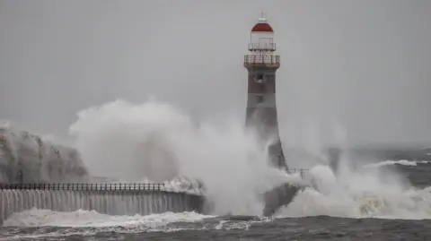 BBC Weather Watchers / Sand Dancer A red and white lighthouse is partially obscured by a crashing wave, which is also cascading over the pier leading up to it. The sky is grey and foggy and the sea also looks grey. There is a large amount of white water spray in the air.