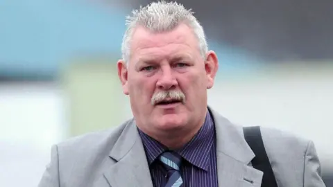 Pacemaker David Tweed is walking looking at the camera. He has short white hair and moustache. He is wearing a grey blazer, purple pin stripped shirt and stripped tie. He is carrying a bag over his shoulder.