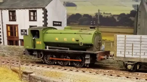 A lime green engine with the spaced out yell letters NCB emblazoned on its side. It is running over a small brick bridge and there is scorched yellow grass to the side. There are two houses in the background close to the track. 