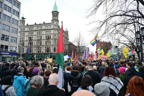 Pacemaker Hundreds of people hold up flags and signs to protest against a mass migration demonstration. They stand on a footpath beside a road. Some of them wear Hi-Viz vests. Signs saying 'people before profit' 'SDLP' and pride flags can be seen, as well as NIPSA flags. 