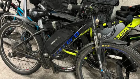 O bicicletă neagră cu inscripție albastră este lipită de alte biciclete. Are un pachet mare de baterii prins de ea cu bandă adezivă gri groasă.