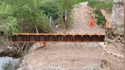 A large metal beam is craned across a river bank while several workers in high-vis watch on.