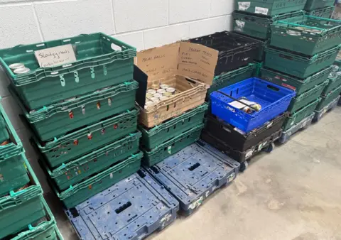 BBC News The photo shows towers of crates piled on top of one another. In them are tinned items, and there are signs indicating the content of them. One reads 'stodgy meals' and another reads 'meatballs and chilli and hot dogs.'