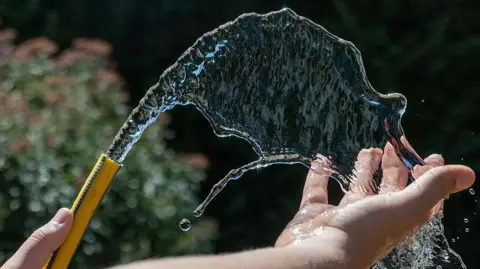 Getty Images A person using a yellow garden hose which is spraying water into the air and on to someone's hand.