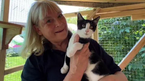 Michele Franks is holding a black and white cat and smiling, she is standing inside an outdoor enclosure of wood and wire construction. 