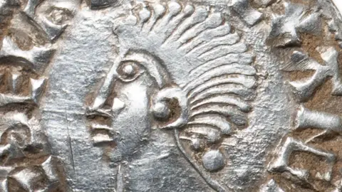 A close up of a silver penny of Ethelred II, king of England. It shows a profile portrait of the king with a mass of hair like a long Mohican. Letters from his name, struck on the circumference can be seen.