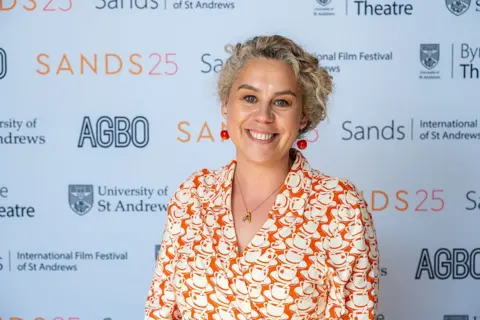Getty Images Ania Trzebiatowska, a blonde, curly-haired woman stands in front of a promotional backdrop for the Sands: International Film Festival of St Andrews wearing a dress printed with orange and white teacups.
