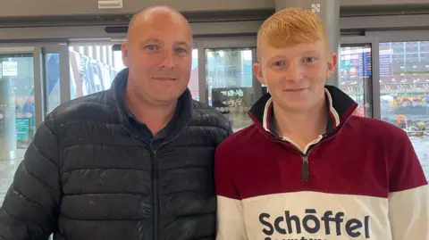 Two men looking at the camera. One is wearing a black jacket the other a red and white quarter zip jumper. He has red hair.