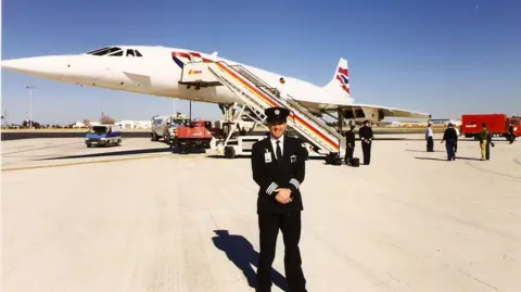 John Tye standing in front of Concorde plane Alpha Foxtrot on the runway. He is dressed in his black pilot uniform and a black cap. He has his hands crossed in front of him. Behind his, others can be seen fixing up the plane, with a bright blue sky above them.