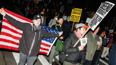 Protesters march along a street in New York City in the dark. A man holds a US flag up behind him as he walks, while another holds a sign saying: "Defend democracy: It's time for a general strike!"