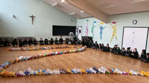 A wider shot of a school hall, children sitting round the edge on the floor. The middle has a twisting stream of cereal boxes.