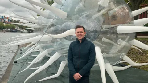 Luke Jerram is stood in front of the giant E.coli next to the water.