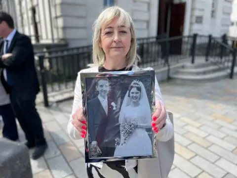 Collette Rooney has light blond hair down to her shoulders and is holding a black and white wedding photograph of her dad and mum while standing outside court  
