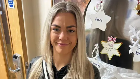 Charlotte pictured with long blonde hair and is wearing black sweatshirt. She is standing by a wall with christmas decorations, coloured in stars by children. 