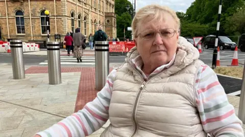 BBC/Seb Cheer A woman, Flick, sits in a wheelchair in front of a large metal bollard, which is sticking up out of a section of red tactile paving. The paving leads to a Zebra crossing, with people walking across it.