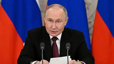 Vladimir Putin, with three Russian flags behind him, holding a piece of paper and speaking into two microphones. 