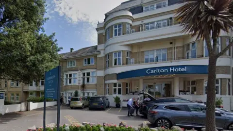 A four-storey art deco style hotel stands behind a small circular flower bed, with several cars parked in front of the building. Large green trees stand on both sides of the building.