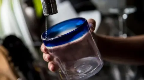 A drinking glass with blue rim being held under a tap with just a small dribble of water coming out