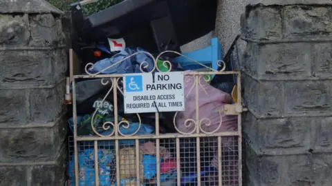 LDRS A mound of rubbish stacked behind a small metal gate which bears a sign saying 'NO PARKING: Disabled access required at all times'.