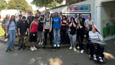 West Northamptonshire Council A large group of students, standing by an underpass, with artwork on the walls. One student to the right is in a wheelchair. 
