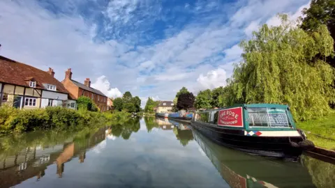 WEDNESDAY - Three narrowboats are moored on the canal bank, the first one is red and green, behind the boats are blue and red. The still water of the canal is flanked on one side by a bank of green grass and a tree, on the other side are houses behind a hedge. The sky overhead is blue with white fluffy clouds that are reflected in the water.