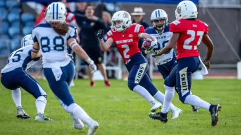 A number of women are playing American football. Two have red tops the others white and blue. A referee is between them in the distance. They are all wearing the heavy protective clothing associated with the game 