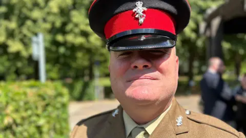 BBC News A man wearing an Army uniform with khaki jacket and light-green shirt and tie, along with a black and red cap with a silver badge. He is standing in the grounds of a crematorium.